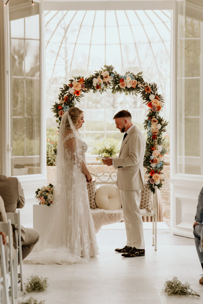 bruidspaar spreekt trouwgeloften naar elkaar uit voor bloemenboog